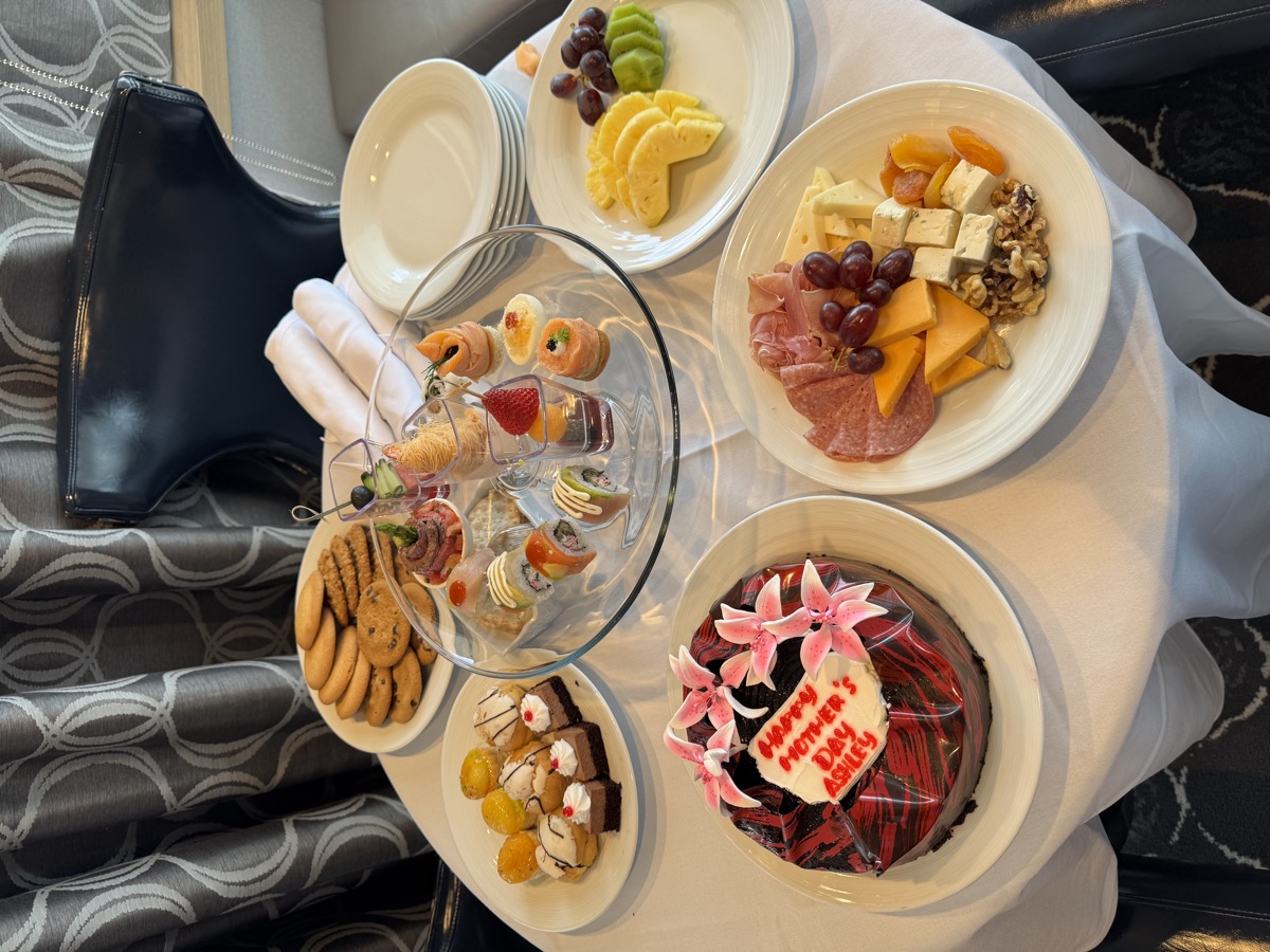 Round table set with charcuterie, fresh fruit, tea sandwiches on a tiered stand, cookies, petit fours, and a chocolate cake reading "Happy Mother's Day Ashley"