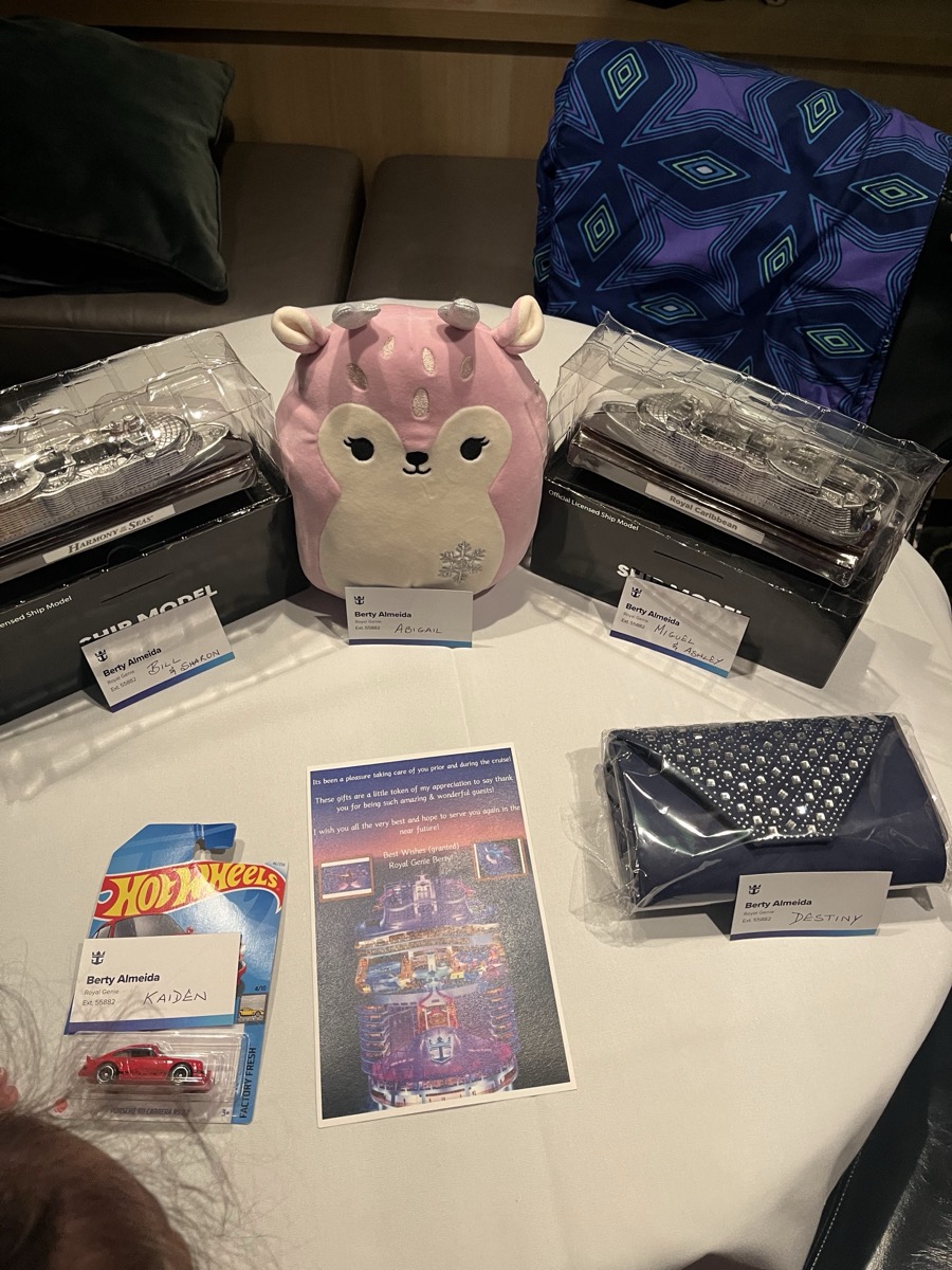 Berty's farewell gifts laid out on a table — Harmony of the Seas ship model, pink Squishmallow, Hot Wheels car, navy beaded clutch, blue scarf, and handwritten note from Royal Genie Berty, with a name placard for every member of the group