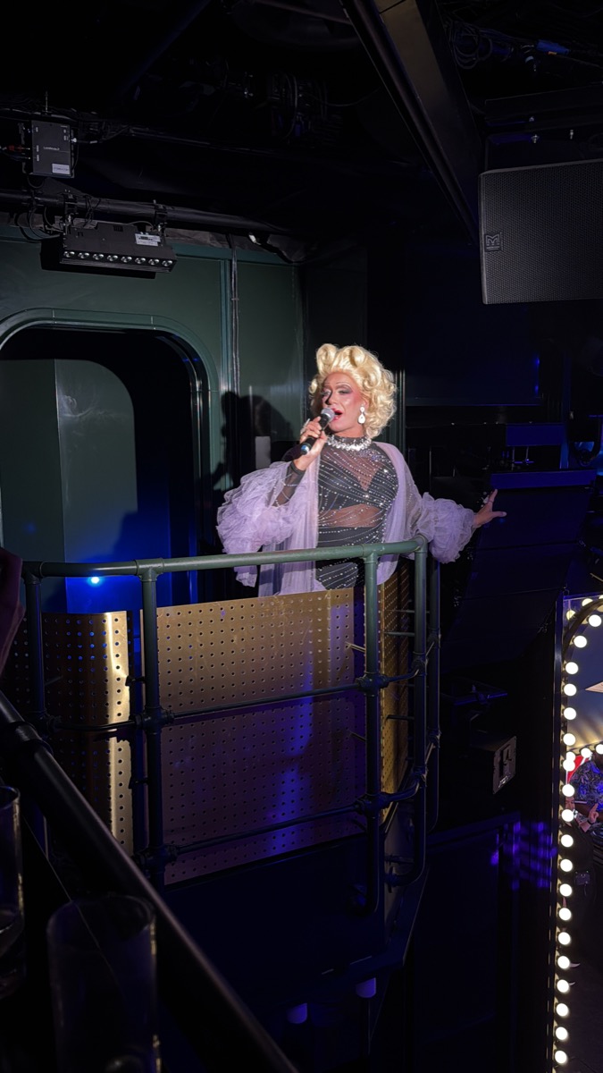 Drag performer in a blonde wig and lavender feathers on the Manor balcony