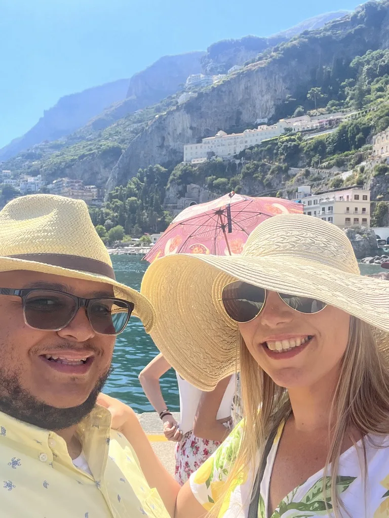 Miguel and Ashley in straw hats taking a selfie at the Amalfi harbor with cliffs behind them
